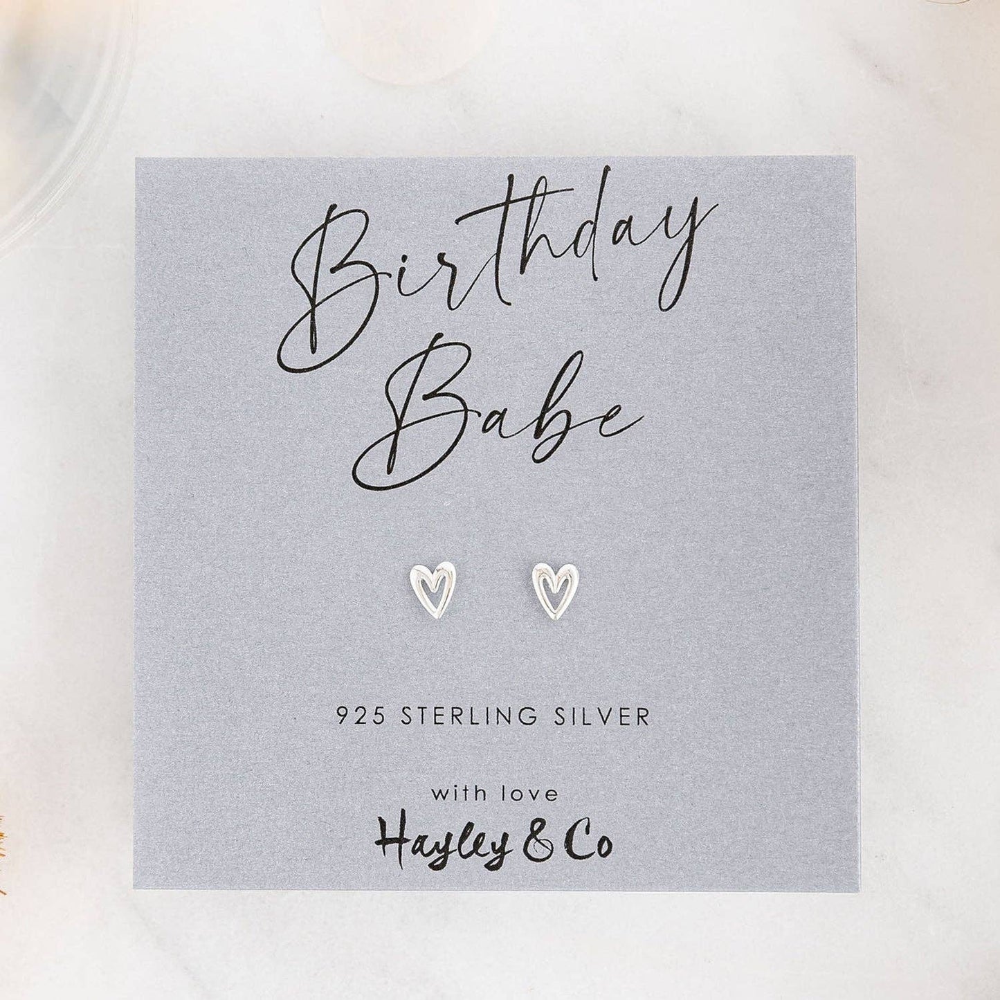 A pair of small silver heart-shaped stud earrings displayed on a gray gift card that reads 'Birthday Babe' and '925 Sterling Silver with love Hayley & Co', photographed on a light marble-like surface.