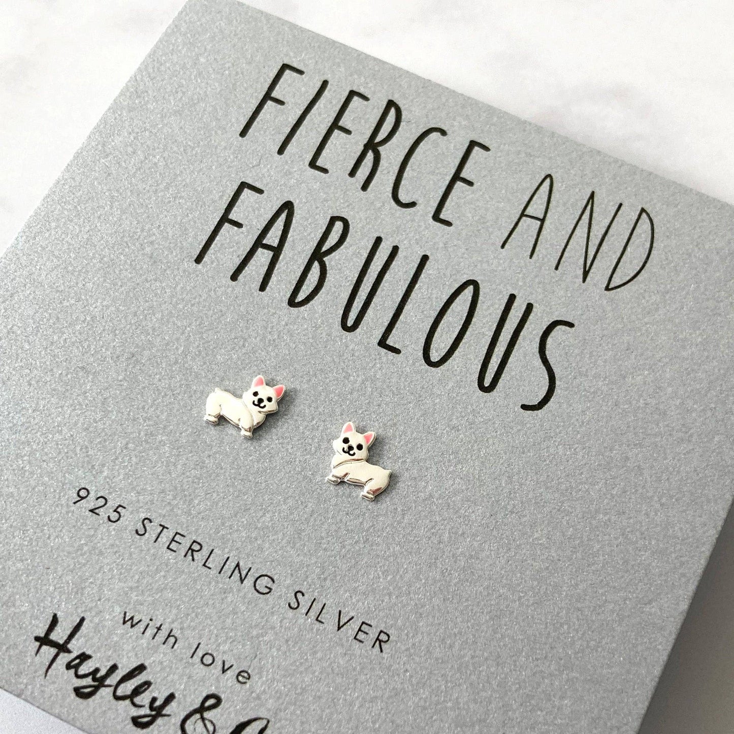 A pair of sterling silver stud earrings shaped like small white dogs with pink ears, displayed on a textured gray gift card that reads 'FIERCE AND FABULOUS' and '925 STERLING SILVER'.