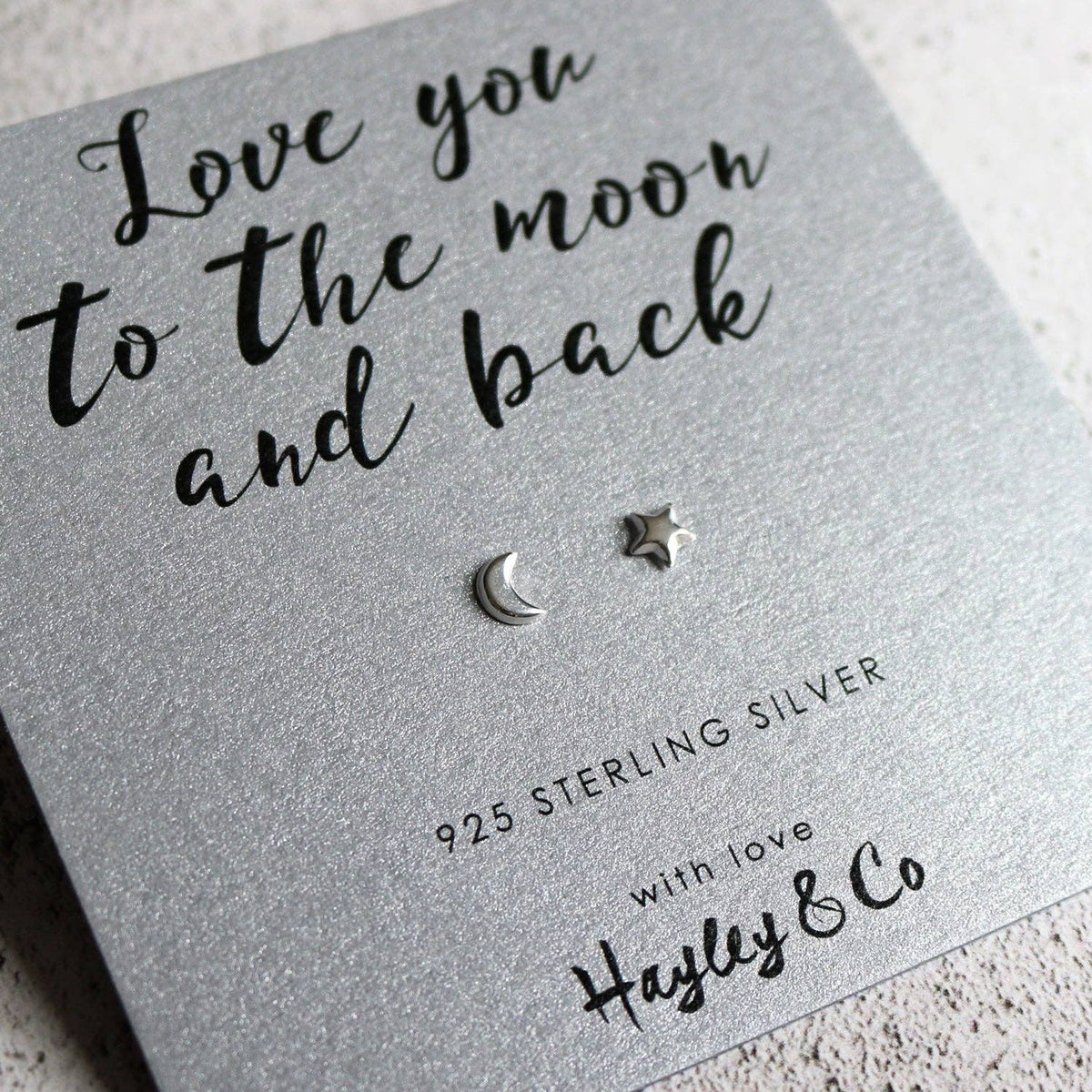 Sterling silver crescent moon and star stud earrings displayed on a textured silver gift card with the phrase 'Love you to the moon and back' and the brand name 'Hayley & Co'.