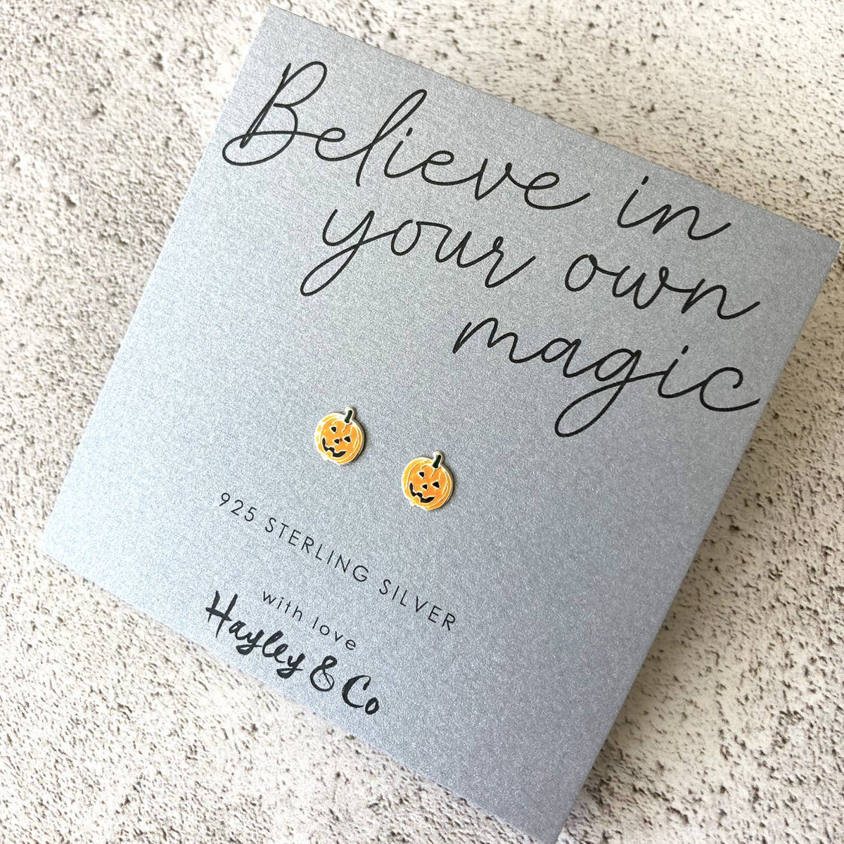 A pair of small pumpkin-shaped stud earrings. Orange with black facial details, displayed on a gray gift card that reads 'Believe in your own magic' and '925 Sterling Silver with love Hayley & Co'.