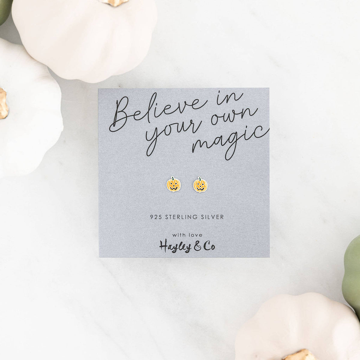 A pair of small pumpkin-shaped stud earrings on a gray display card that reads 'Believe in your own magic' and '925 Sterling Silver with love Hayley & Co,' styled on a white marble surface with decorative pumpkins around it.