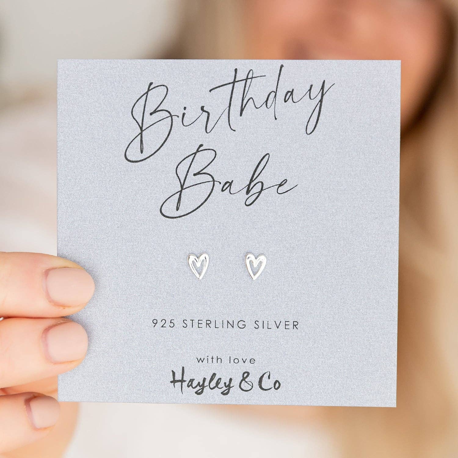 A pair of small silver heart stud earrings displayed on a light gray card that reads 'Birthday Babe' and '925 STERLING SILVER with love Hayley & Co.' A hand holds the card, with a softly blurred person in the background.