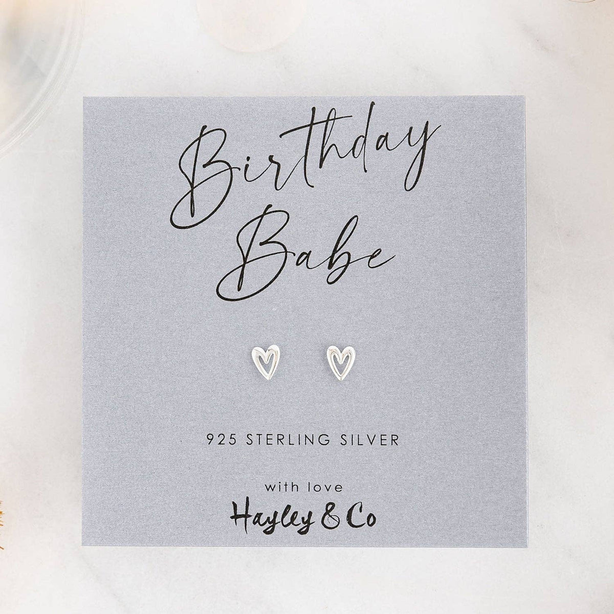 A pair of small silver heart-shaped stud earrings displayed on a gray gift card that reads 'Birthday Babe' and '925 Sterling Silver with love Hayley & Co', photographed on a light marble-like surface.
