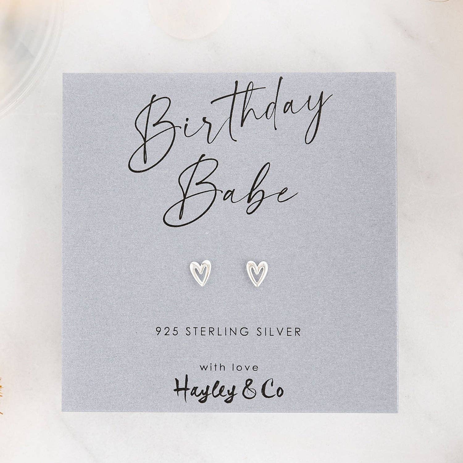 A pair of small silver heart-shaped stud earrings displayed on a gray gift card that reads 'Birthday Babe' and '925 Sterling Silver with love Hayley & Co', photographed on a light marble-like surface.