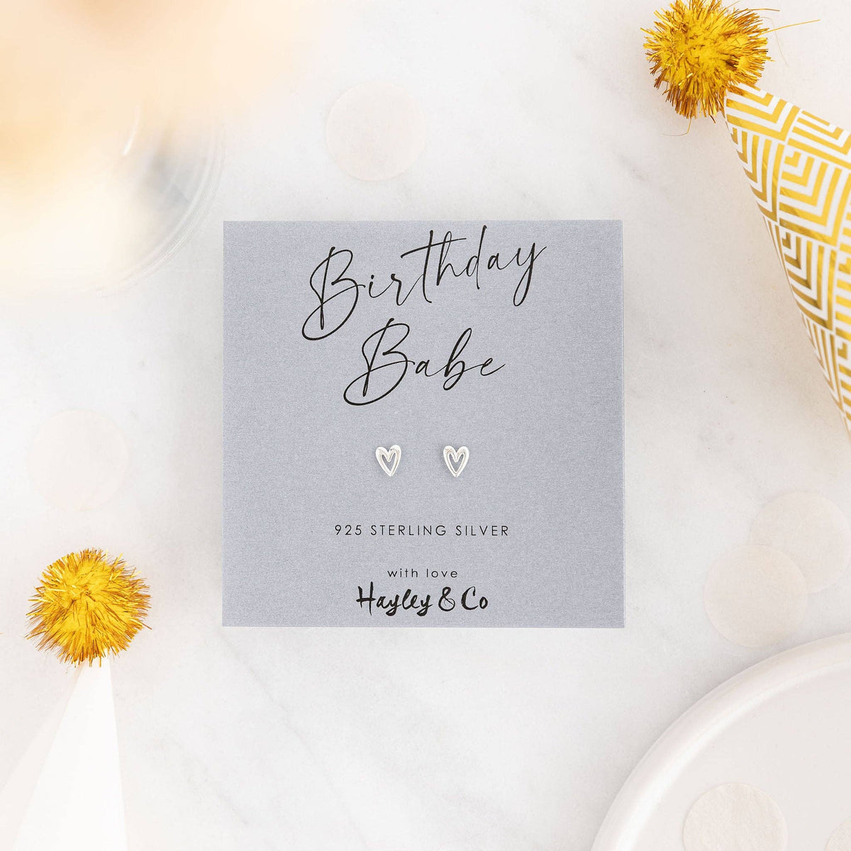 A pair of small silver heart stud earrings displayed on a gray card that reads 'Birthday Babe' with '925 Sterling Silver' and the brand 'Hayley & Co'. The card is styled on a white marble surface with yellow pom-pom flowers and party hats.