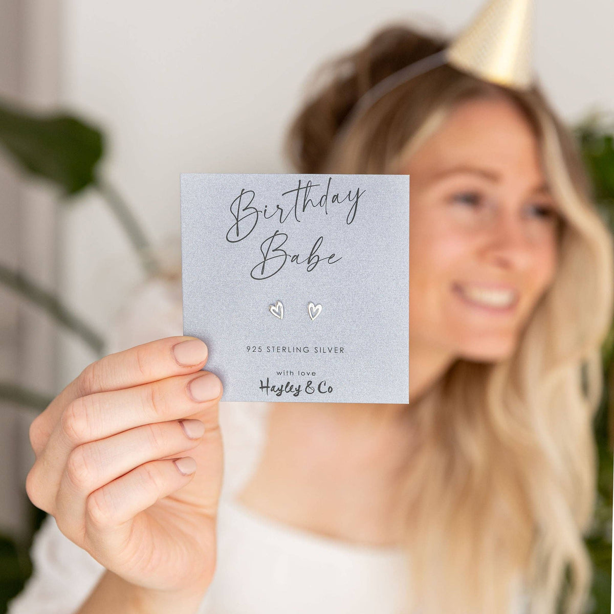 A woman in a party hat holds up a small silver pair of stud earrings on a grey backing card that reads 'Birthday Babe' and '925 Sterling Silver with love Hayley & Co.'.