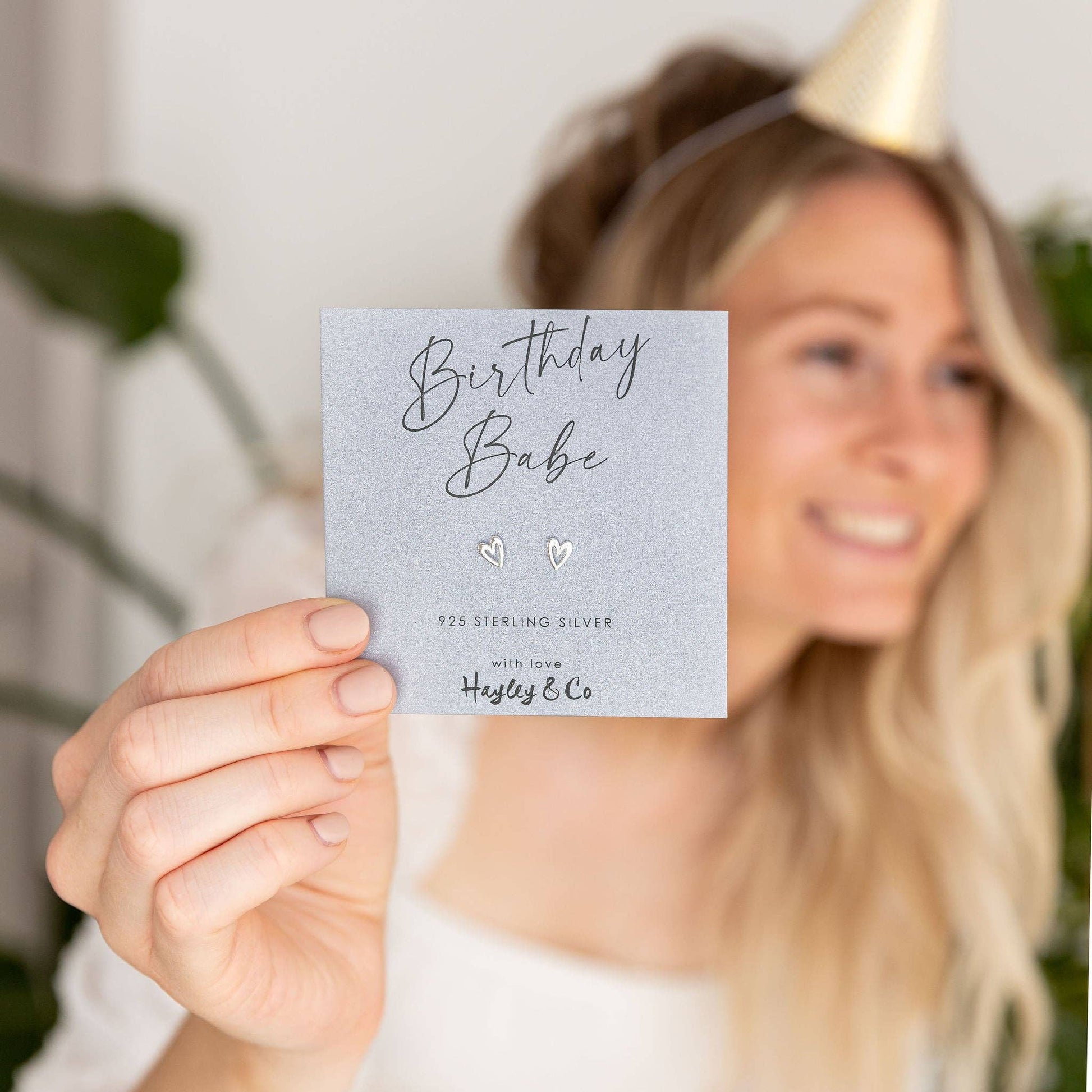 A woman in a party hat holds up a small silver pair of stud earrings on a grey backing card that reads 'Birthday Babe' and '925 Sterling Silver with love Hayley & Co.'.