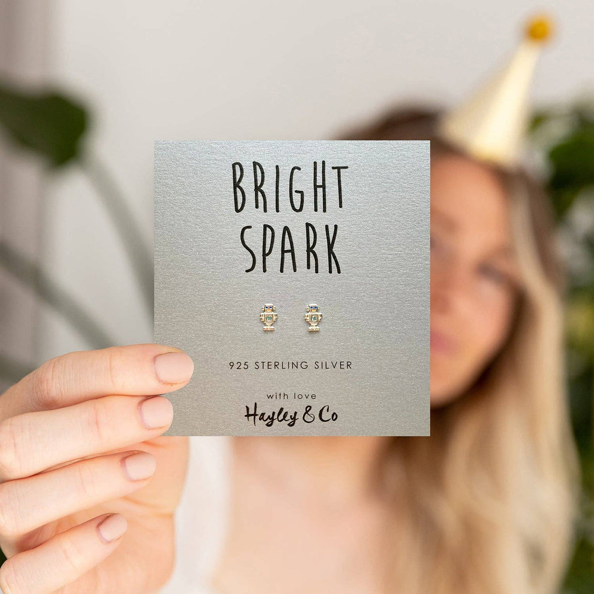 A person holds a silver display card featuring a pair of small silver stud earrings with diamante stones. The card reads 'BRIGHT SPARK' and '925 STERLING SILVER with love Hayley & Co.