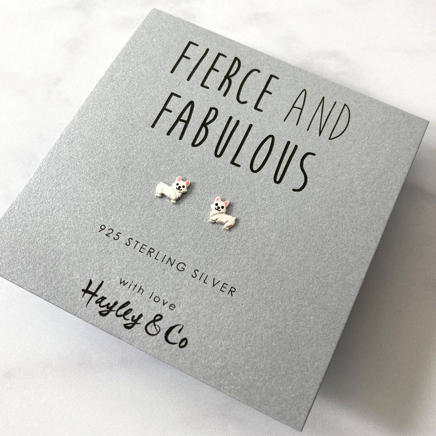 A pair of small dog stud earrings displayed on a textured gray gift card that reads 'FIERCE AND FABULOUS' with 'Hayley & Co' at the bottom. The card is photographed on a light marble-like surface.