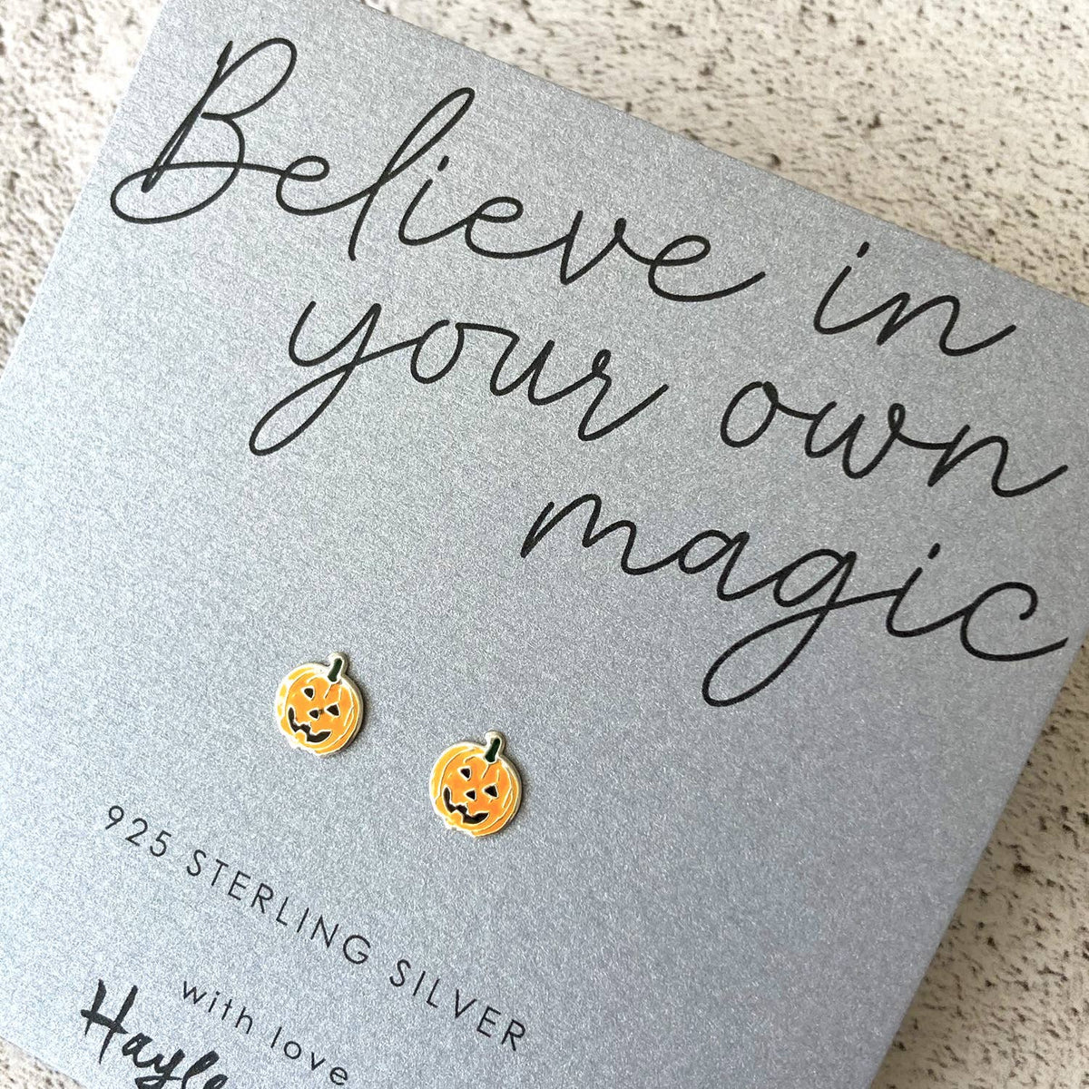A pair of small pumpkin-shaped stud earrings in sterling silver with an enamel pumpkin picture which is orange with black facial details, displayed on a gray gift card that reads 'Believe in your own magic' and '925 Sterling Silver.'.