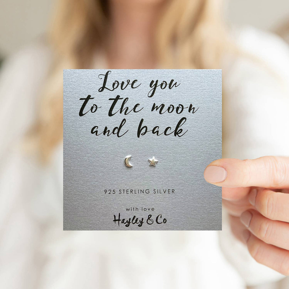 A hand holds a silver gift card featuring a small sterling silver crescent moon and star stud earring. The card reads 'Love you to the moon and back', '925 STERLING SILVER', and 'with love Hayley & Co'.