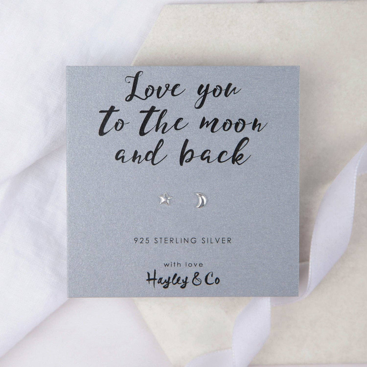 A pair of small silver moon and star stud earrings displayed on a grey gift card that reads 'Love you to the moon and back' with the brand name 'Hayley & Co', set against a soft white and beige fabric background.