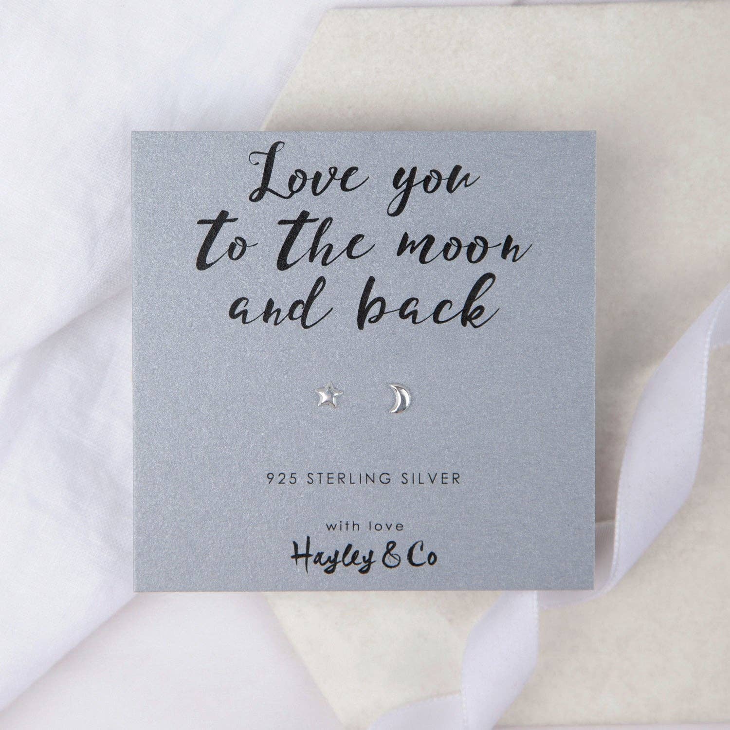 A pair of small silver moon and star stud earrings displayed on a grey gift card that reads 'Love you to the moon and back' with the brand name 'Hayley & Co', set against a soft white and beige fabric background.