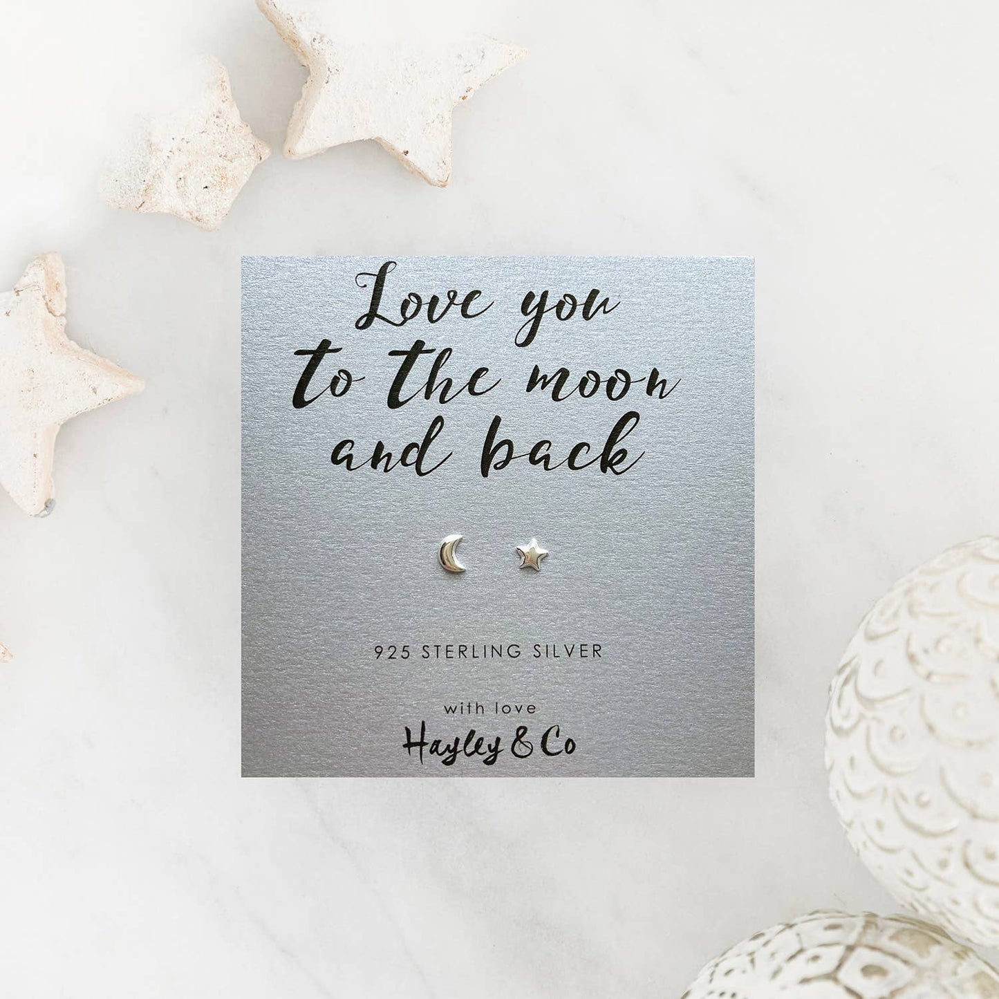 A pair of sterling silver crescent moon and star stud earrings displayed on a silver card that reads 'Love you to the moon and back' with the brand 'Hayley & Co'. The card is styled on a white marble surface with decorative star-shaped and textured ceramic objects.