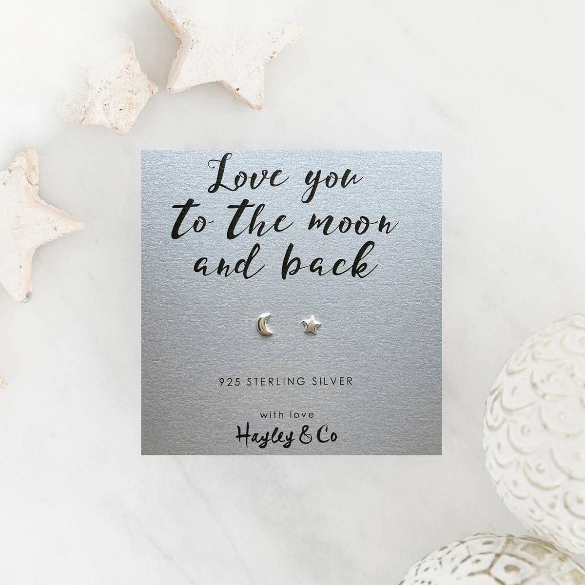 A pair of sterling silver crescent moon and star stud earrings displayed on a silver card that reads 'Love you to the moon and back' with the brand 'Hayley & Co'. The card is styled on a white marble surface with decorative star-shaped and textured ceramic objects.