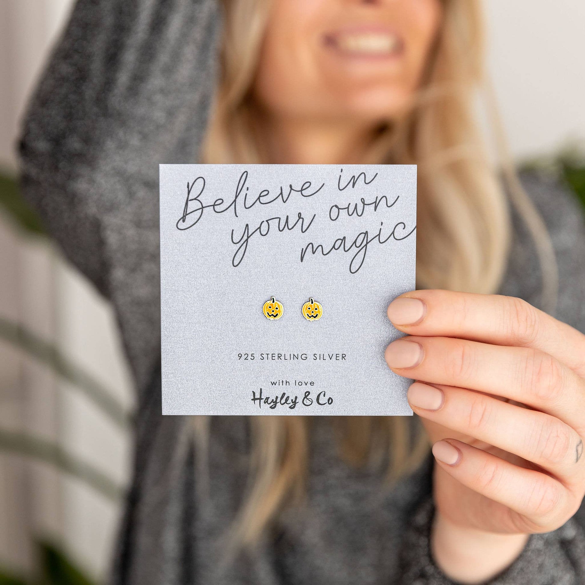 A woman holds a grey jewelry card featuring two small yellow pumpkin (jack-o'-lantern) stud earrings. The card reads 'Believe in your own magic' and '925 STERLING SILVER with love Hayley & Co'.