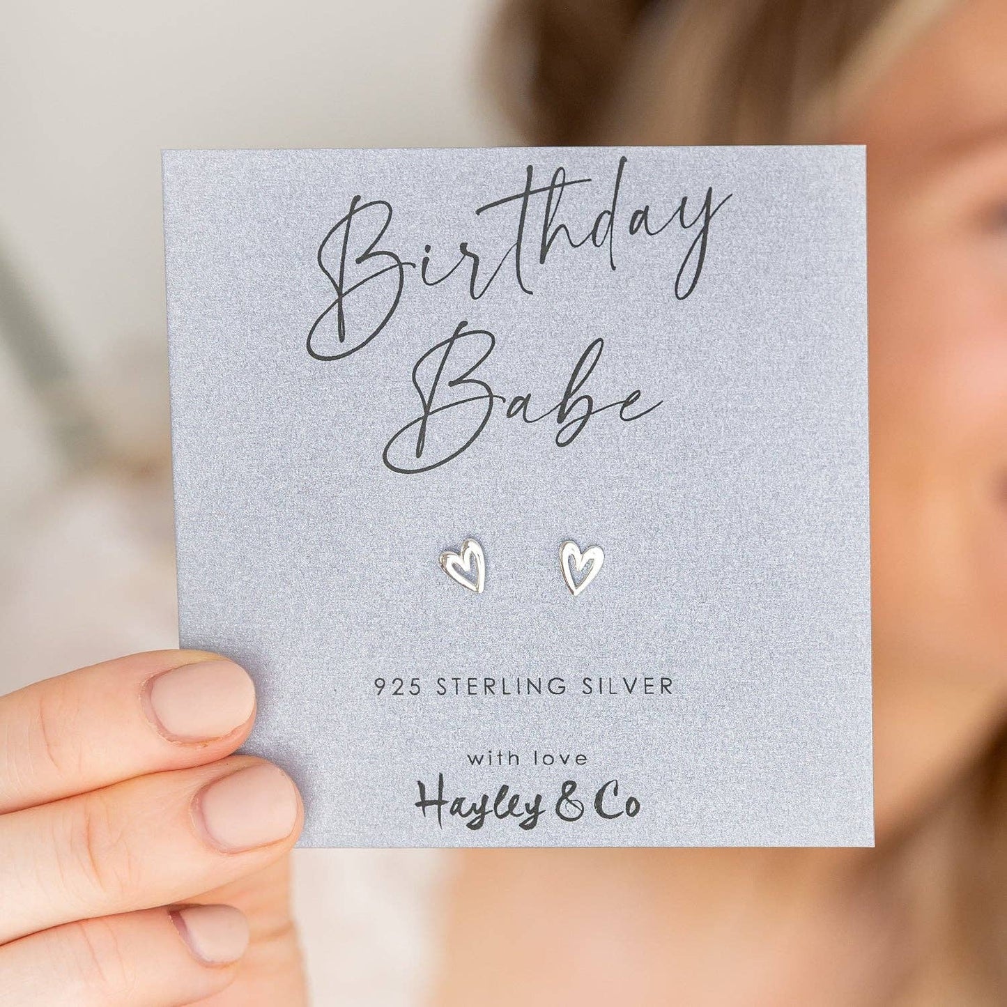 A close-up of a hand holding a silver card with two small heart-shaped sterling silver stud earrings. The card reads 'Birthday Babe' and '925 STERLING SILVER with love Hayley & Co'.