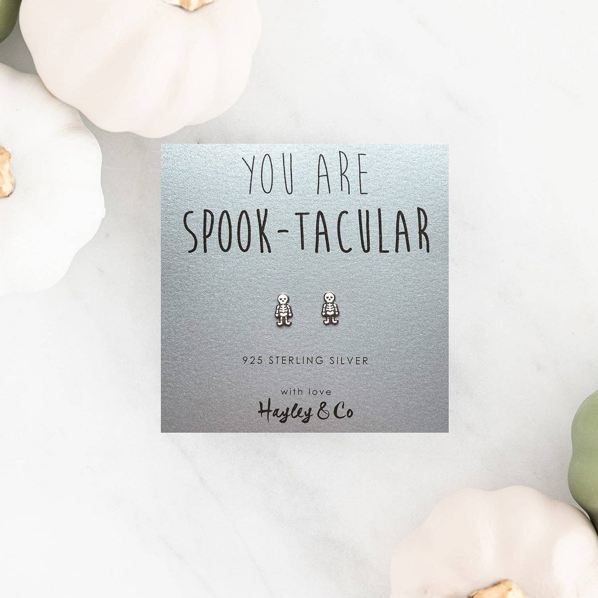 A pair of small silver spook character stud earrings displayed on a square silver card that reads 'YOU ARE SPOOK-TACULAR' with '925 STERLING SILVER' and the brand 'Hayley & Co'. The card is styled on a light marble surface with decorative pumpkins around it.