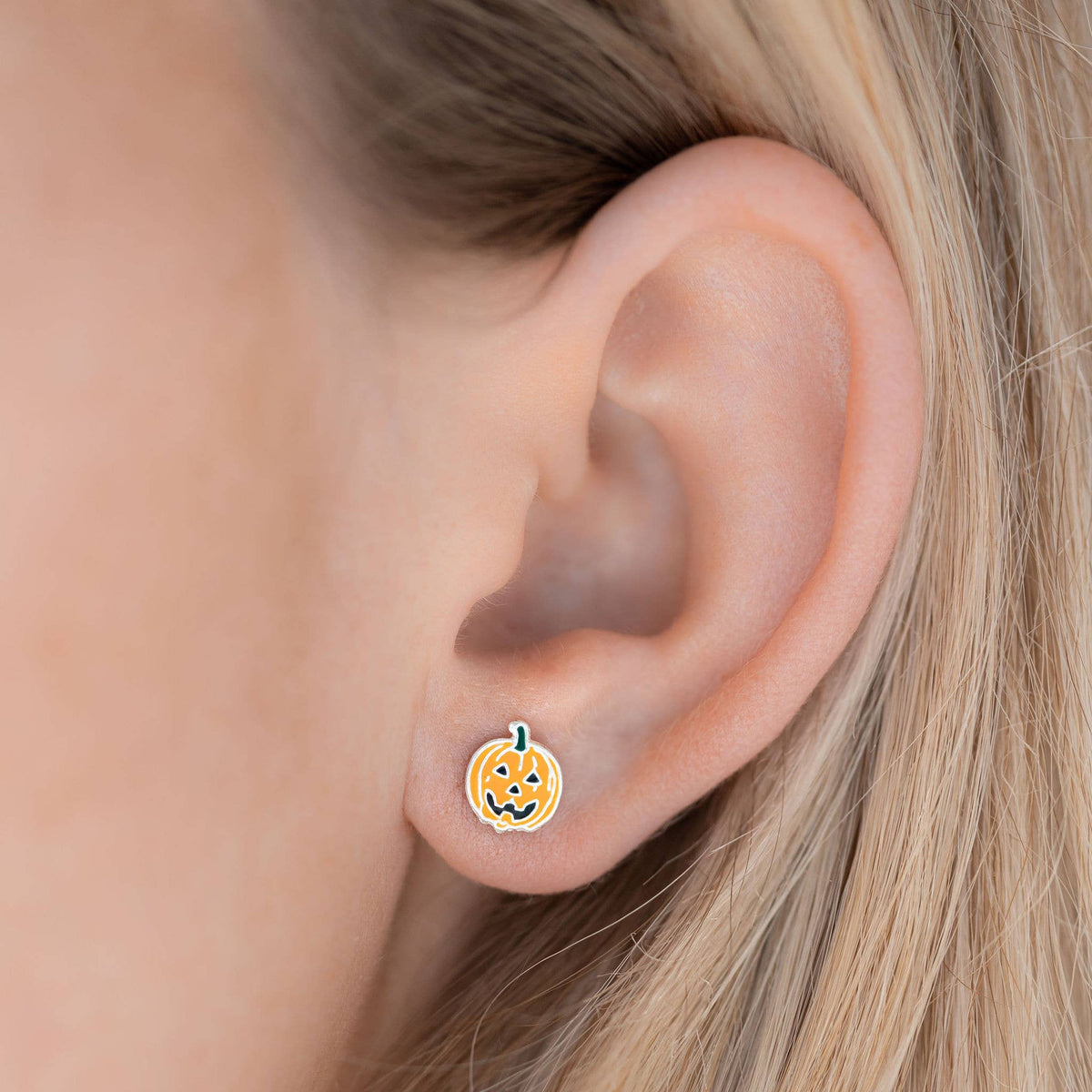 A close-up of a person's ear wearing a small pumpkin-shaped stud earring with a smiling jack-o'-lantern face and a tiny green stem.