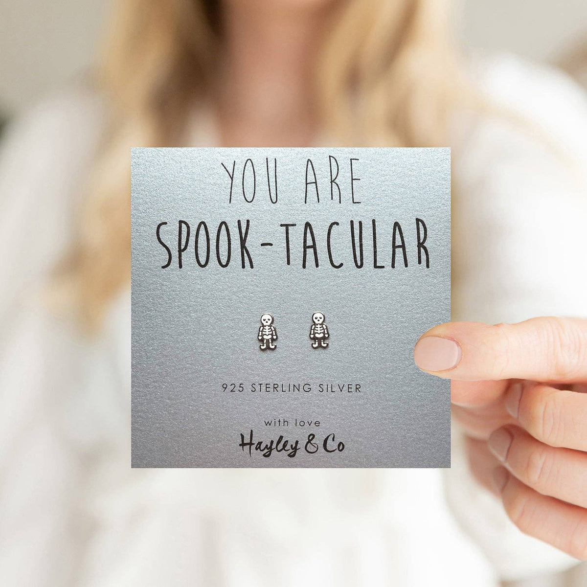 A hand holds a silver gift card featuring a pair of small silver spooktacular character stud earrings. The card reads 'YOU ARE SPOOK-TACULAR', '925 STERLING SILVER', and 'with love Hayley & Co'.